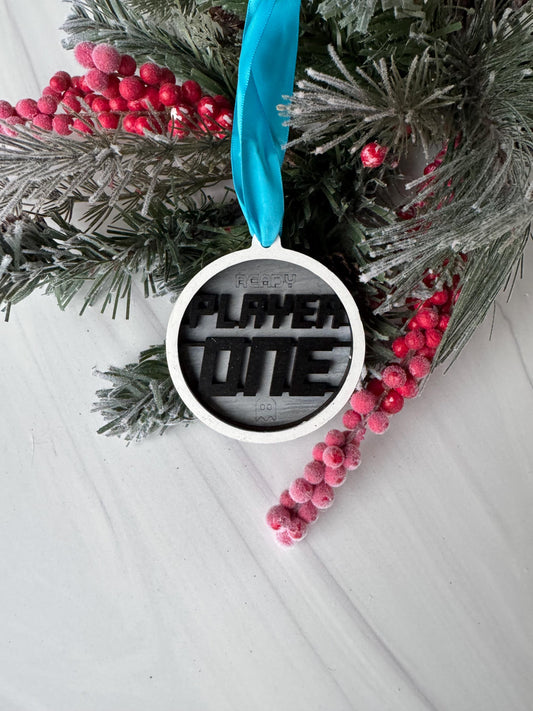 A Christmas ornament with the text &quot;FLY ME&quot; is displayed on a white surface, surrounded by pine branches, red berries, and a blue ribbon.