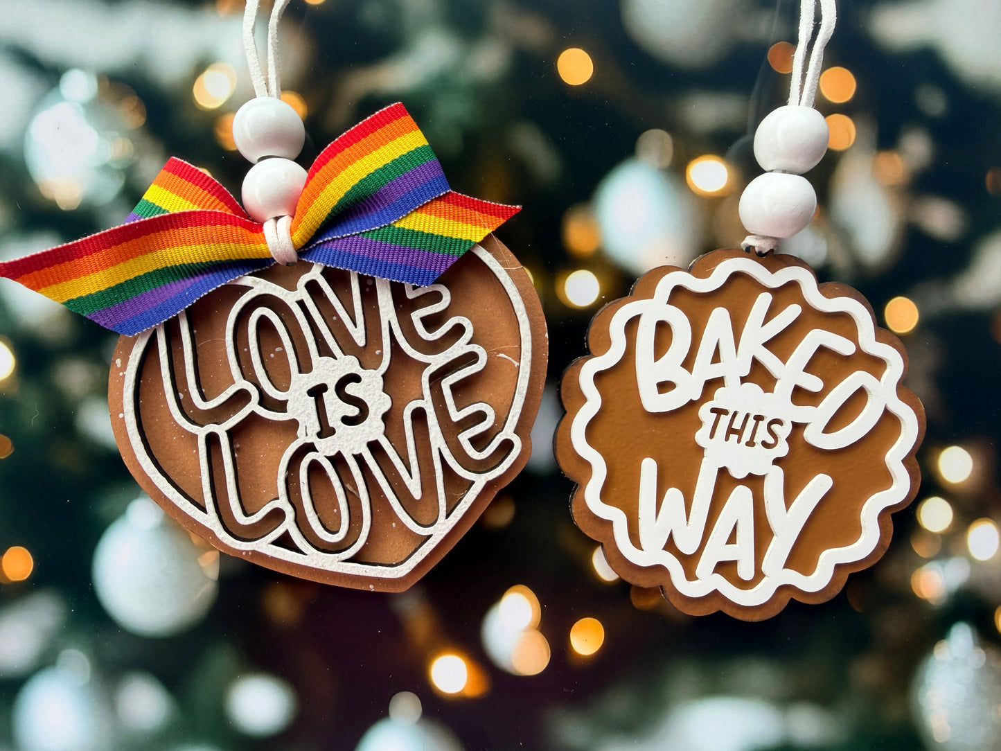 two heart-shaped ornaments hanging from strings, each with a different message. The first ornament has the words "LOVE IS LOVE" written on it, and the second ornament has the words "BAKED THIS WAY" written on it.