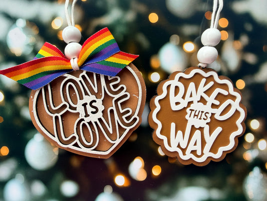 two heart-shaped ornaments hanging from strings, each with a different message. The first ornament has the words "LOVE IS LOVE" written on it, and the second ornament has the words "BAKED THIS WAY" written on it.