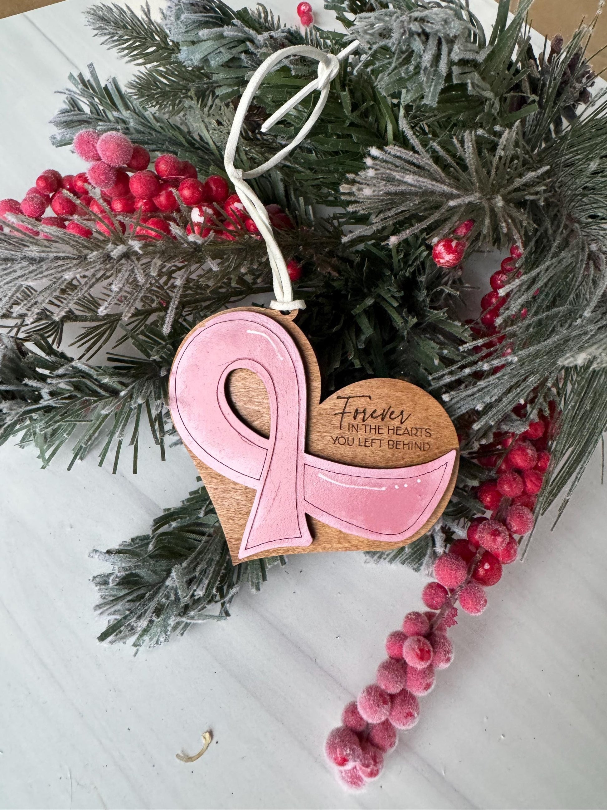 A pink ribbon ornament is displayed on a pine branch, surrounded by red berries and greenery, with a white background.