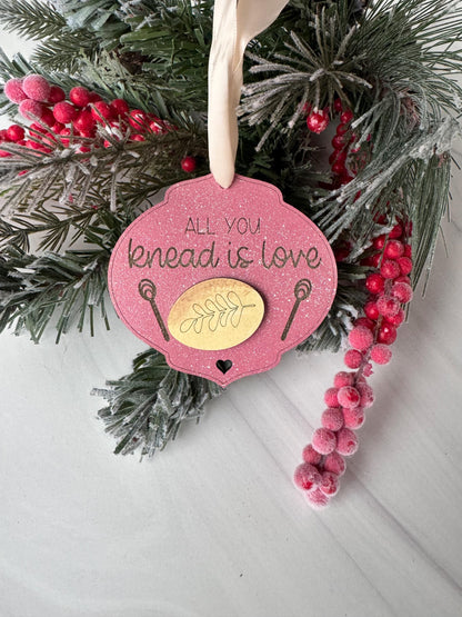 A pink ornament with the text "All you need is love" is surrounded by pine branches, red berries, and a white ribbon.