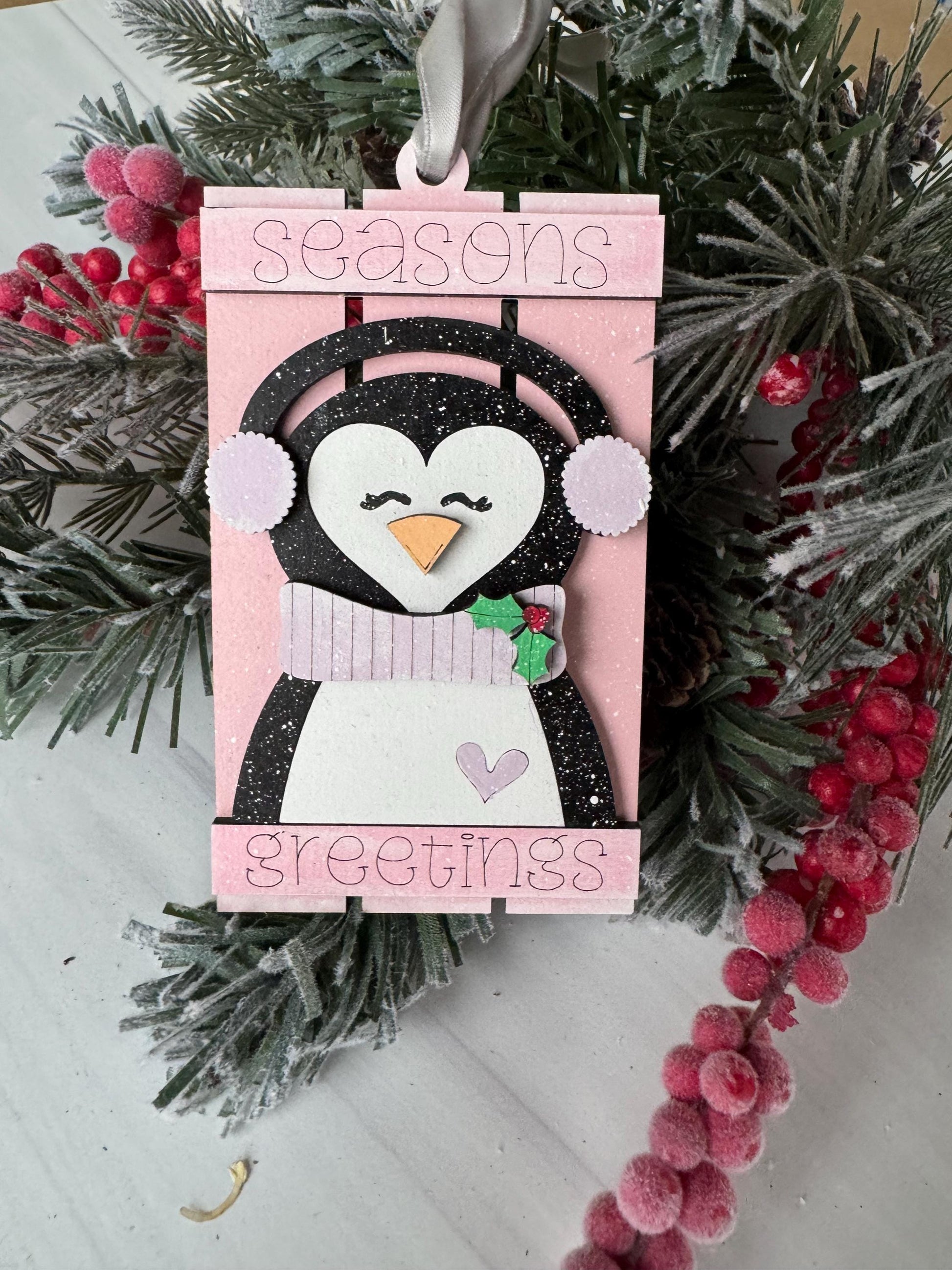 A pink ornament with a penguin design, surrounded by a wreath of berries and pine branches, with the word "Seasons" written on it.