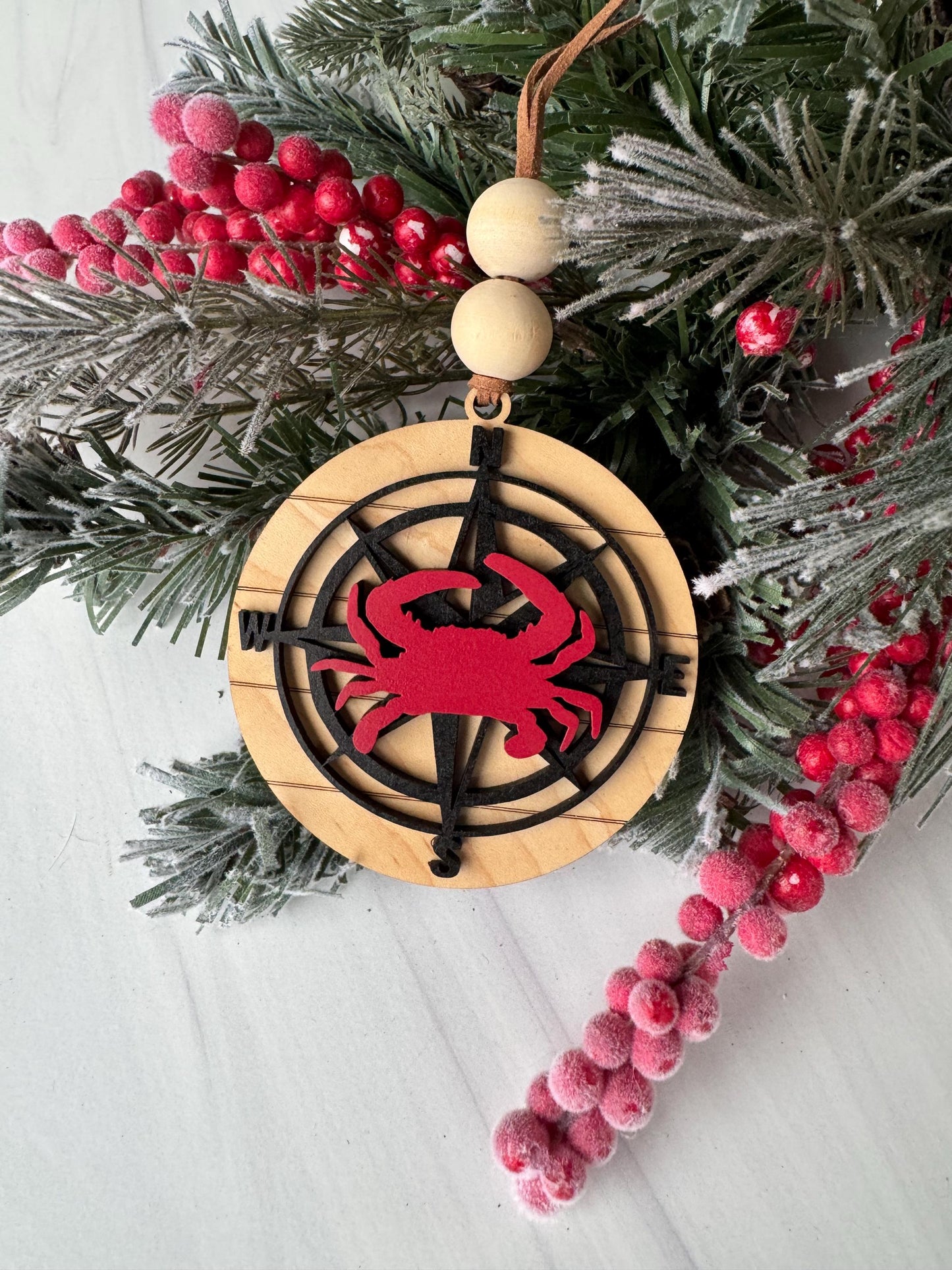 A Christmas ornament with a red crab design on a wooden background, surrounded by pine branches, berries, and a compass rose.