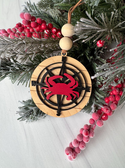 A Christmas ornament with a red crab design on a wooden background, surrounded by pine branches, berries, and a compass rose.