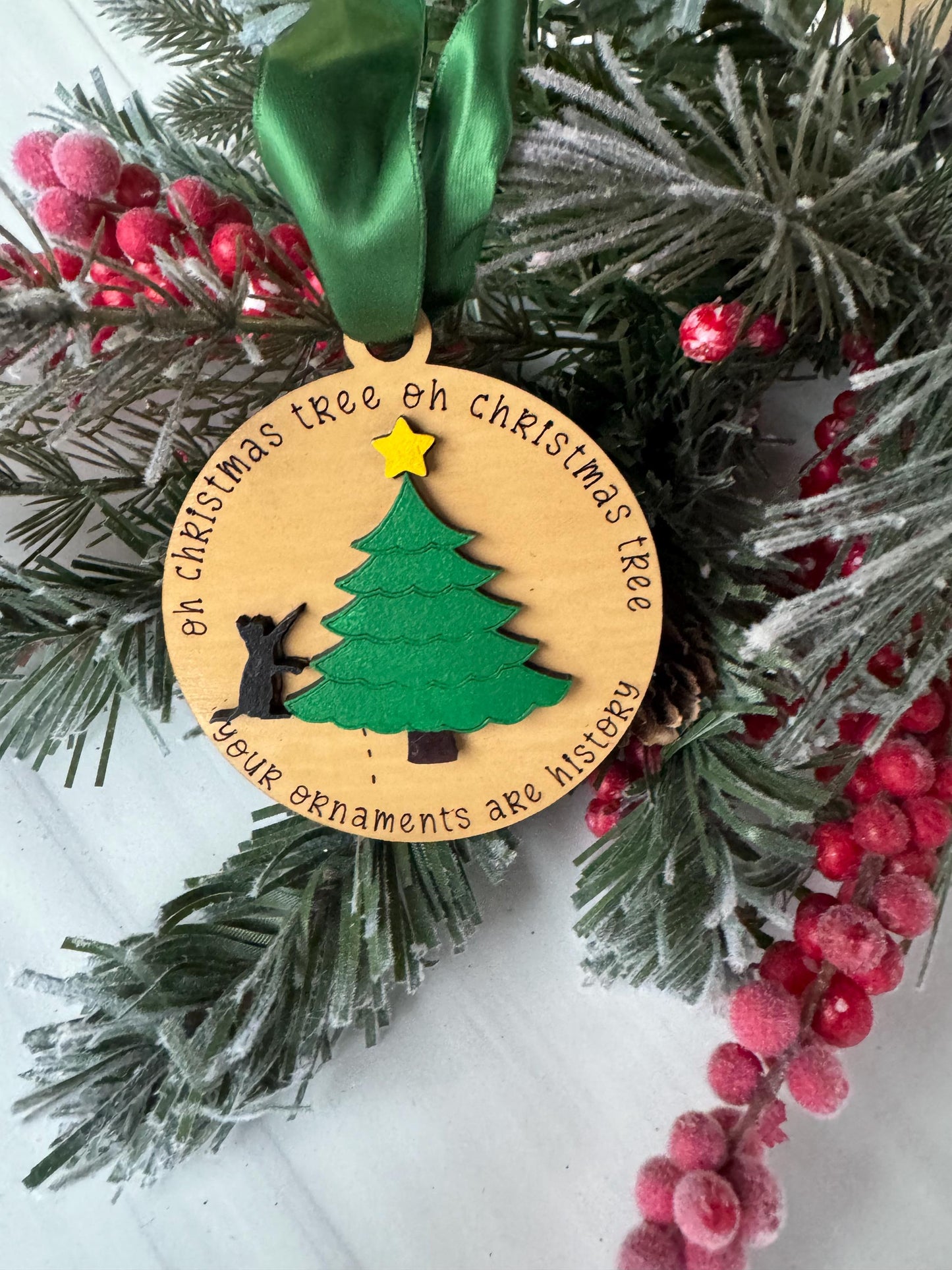 A wooden ornament with a Christmas tree design, surrounded by pine branches, red berries, and a green ribbon.