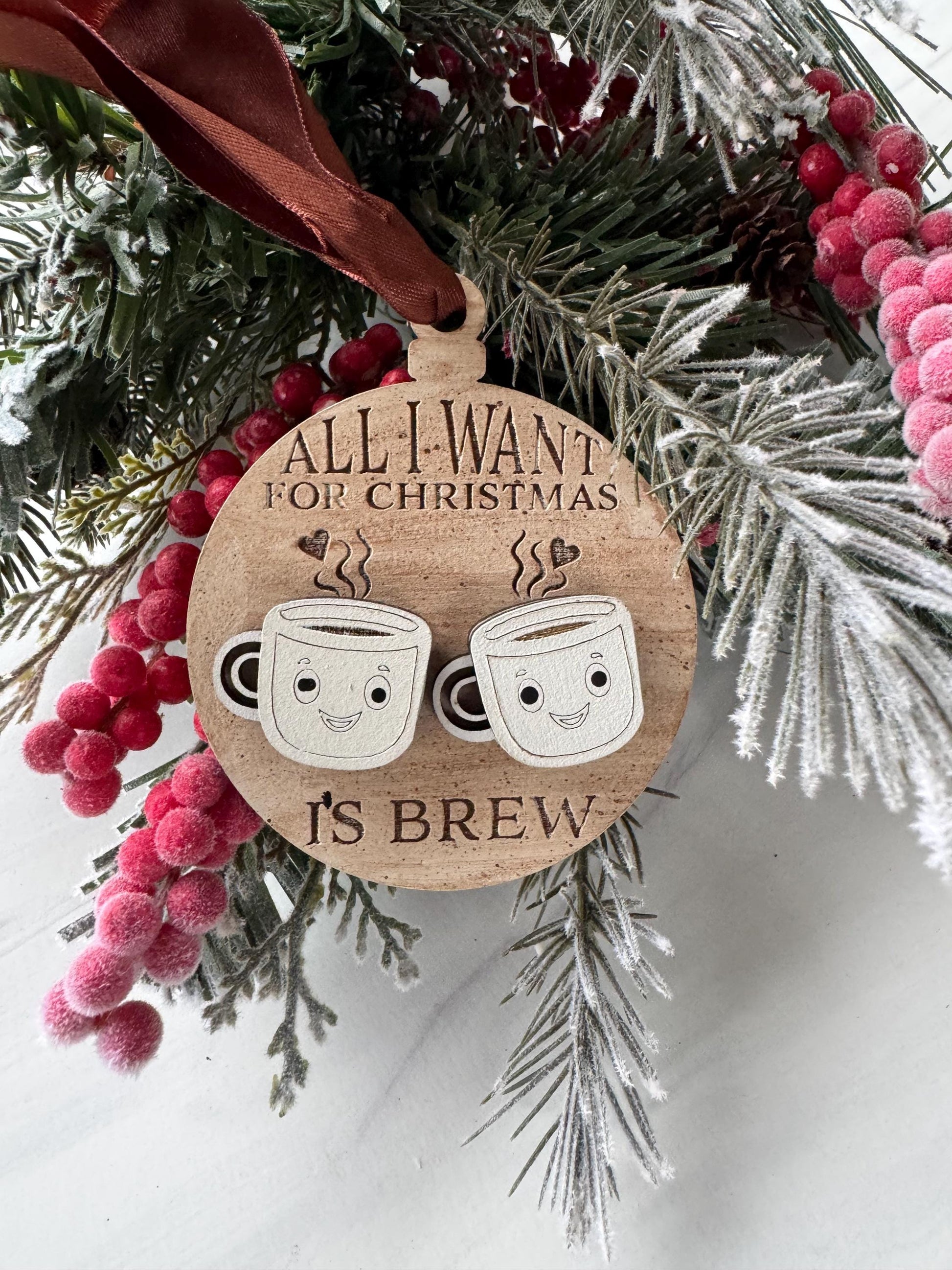 A wooden ornament with a wreath of pine branches and red berries, featuring a pair of cartoon coffee cups and the text "All I want for Christmas is coffee".