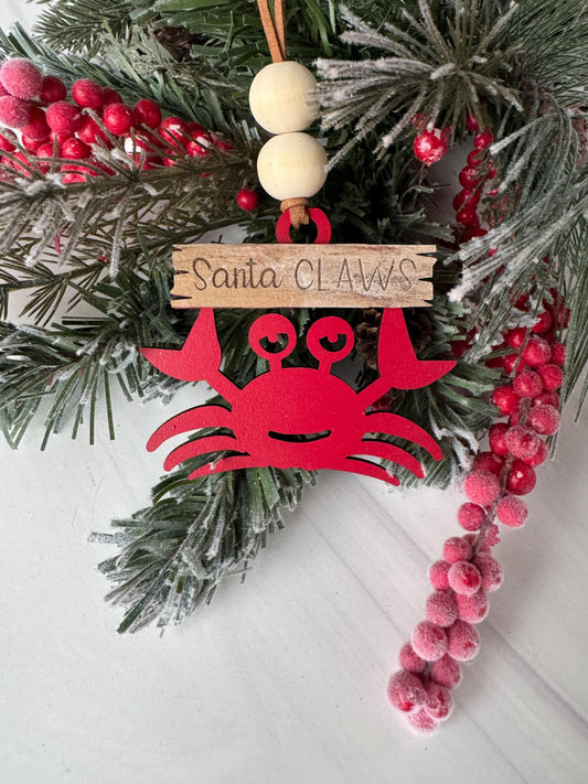 A red ornament resembling a crab is hanging on a Christmas tree, surrounded by pine branches, berries, and a wooden sign.