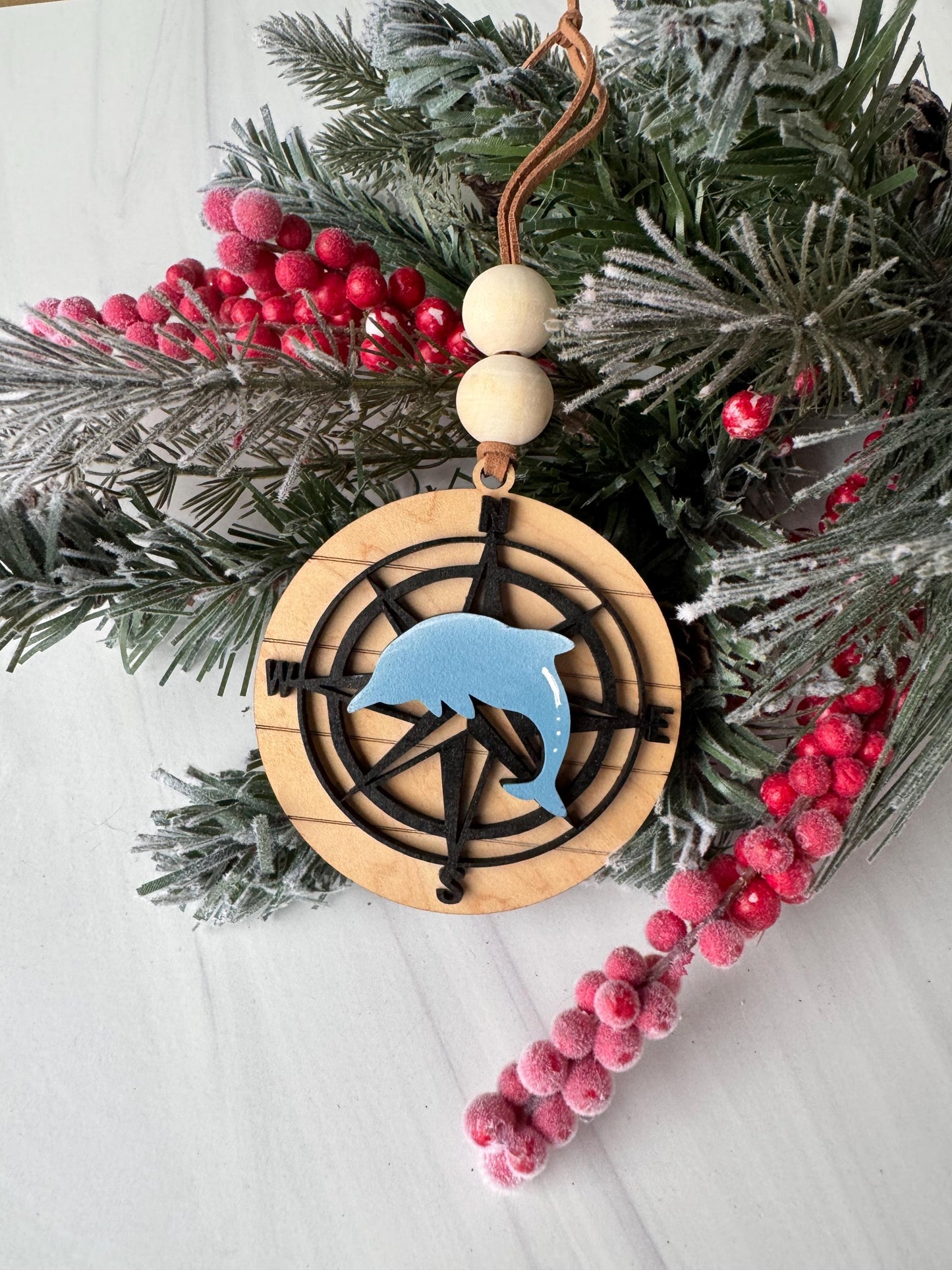 A wooden compass ornament with a blue dolphin on a white background, surrounded by a wreath of pine branches and red berries.