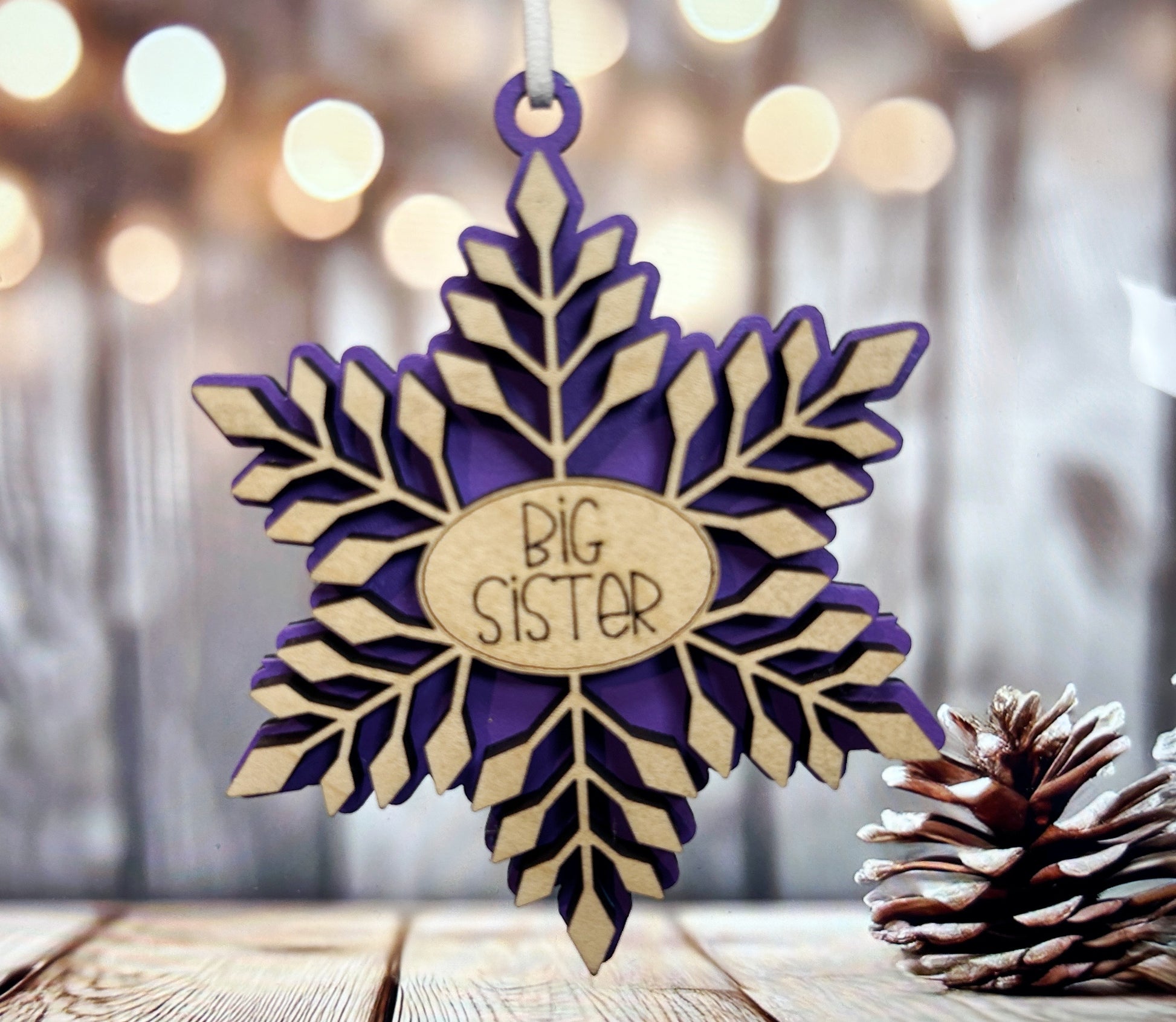 A purple snowflake ornament with the text "BIG SISTER" is hanging from a string, with a pine cone and blurred background lights visible.