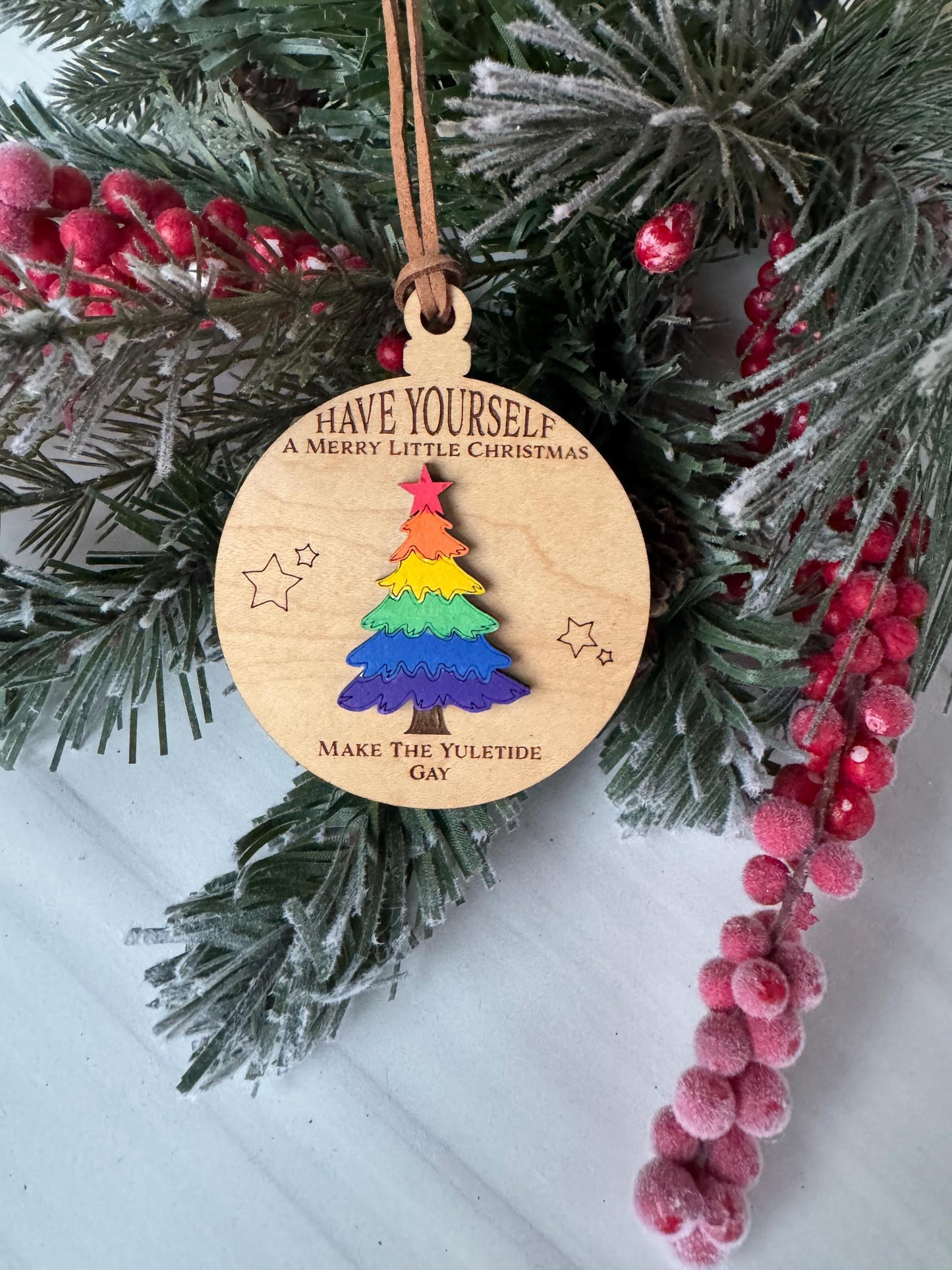 A wooden ornament with a colorful tree design hangs on a Christmas tree branch, surrounded by pine branches and red berries.