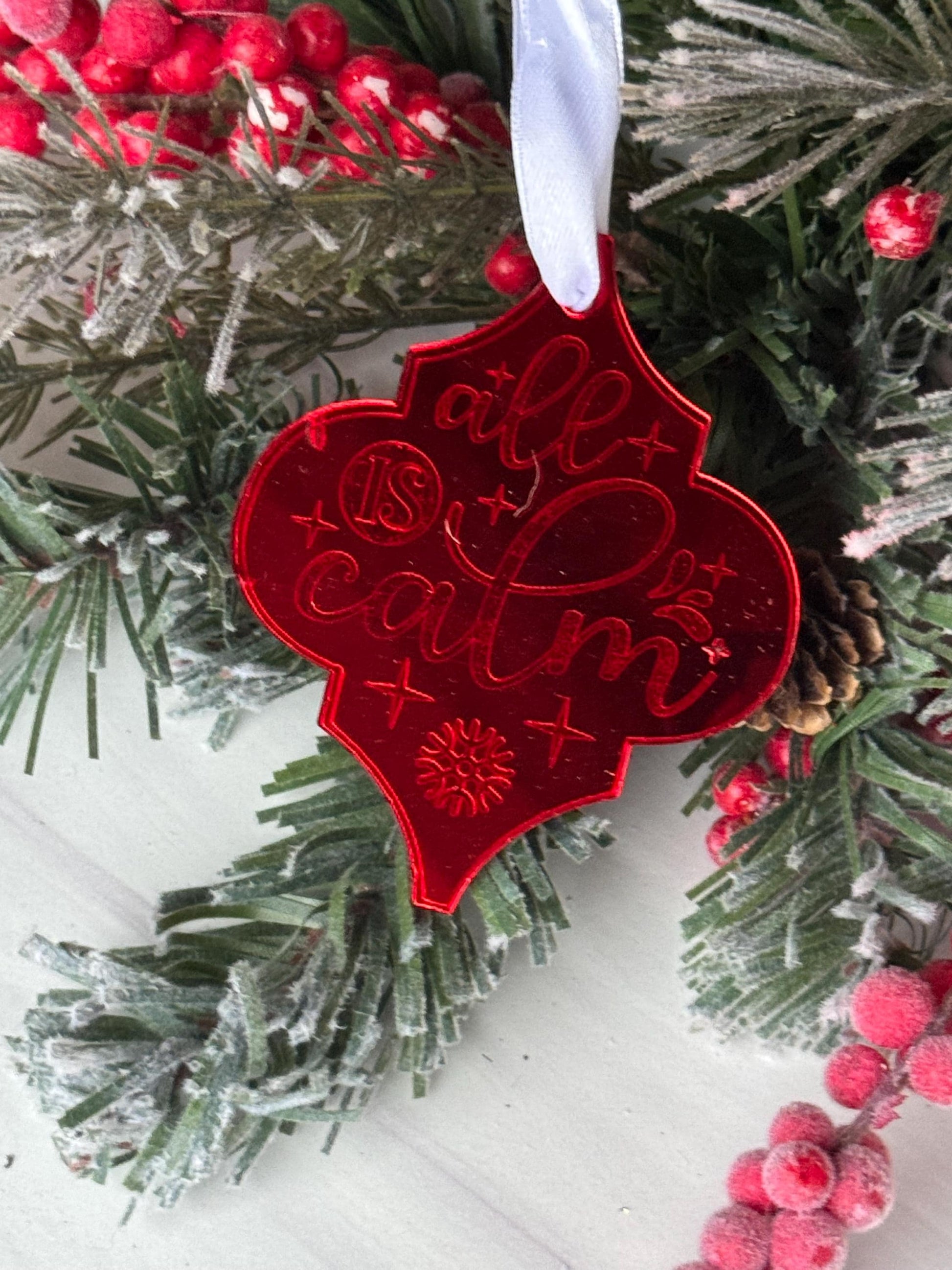 A red ornament with a floral design is displayed among pine branches and red berries, creating a festive holiday scene.