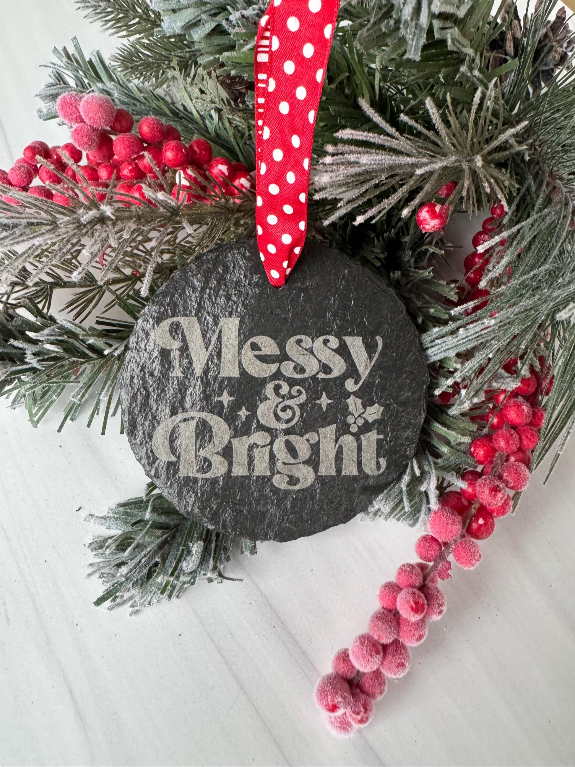 A black stone ornament with the text "MESSY & BRIGHT" is surrounded by a wreath of pine branches, red berries, and a red polka dot ribbon.