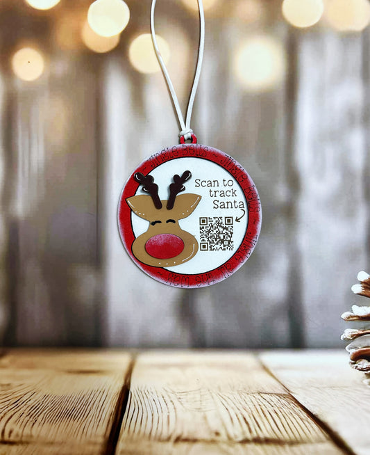 A Christmas ornament with a reindeer and the text "Scan to track Santa" is hanging on a string against a blurred background with bokeh lights.