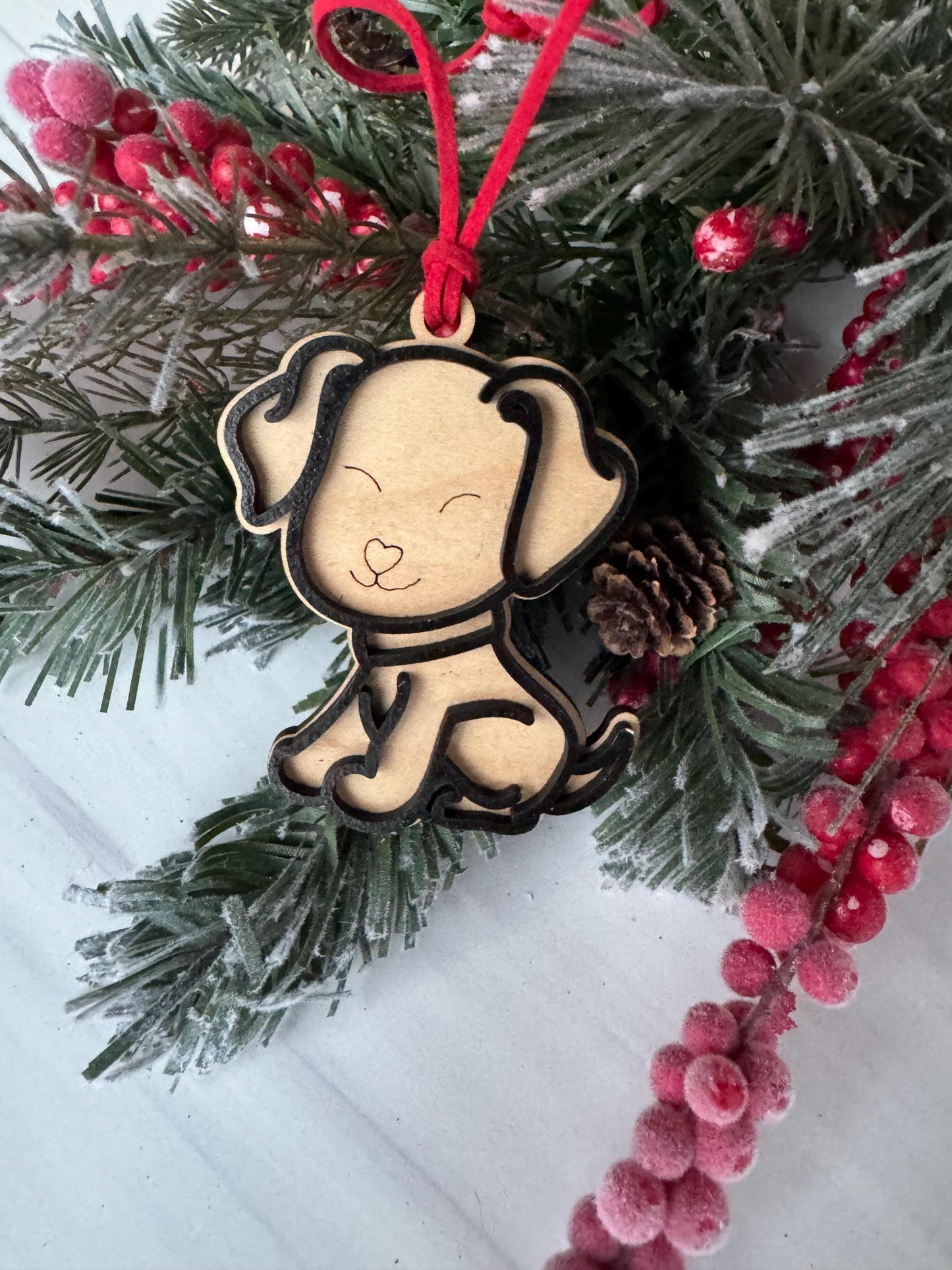 A wooden ornament in the shape of a dog is hanging on a Christmas tree, surrounded by pine branches and red berries.