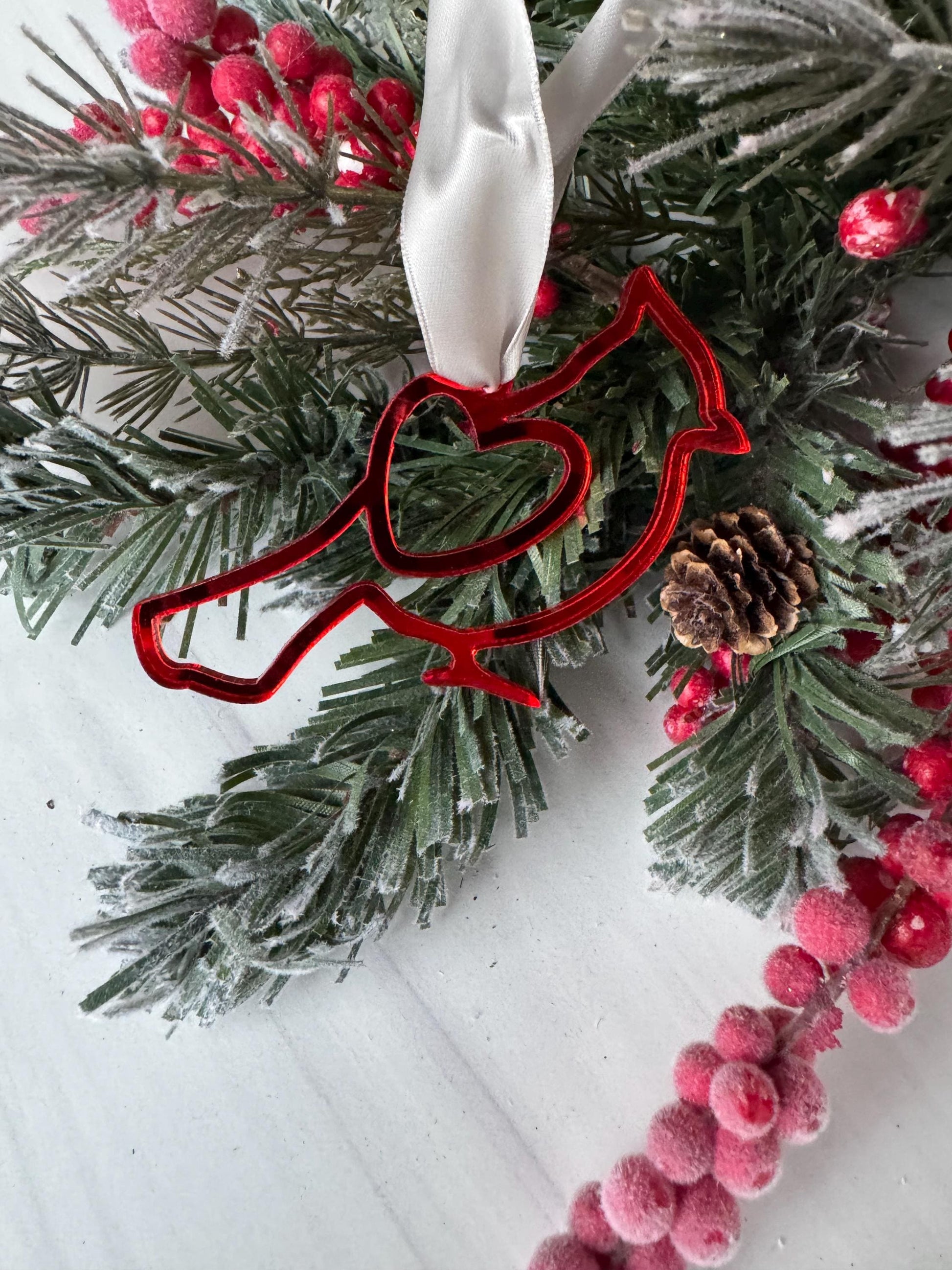 A red bird ornament is hanging from a branch of a Christmas tree, surrounded by pine needles, berries, and a white ribbon.