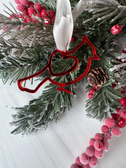 A red bird ornament is hanging from a branch of a Christmas tree, surrounded by pine needles, berries, and a white ribbon.