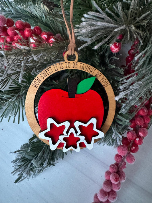 A wooden ornament with a red apple and three white stars is displayed on a Christmas tree branch, surrounded by pine branches and red berries.