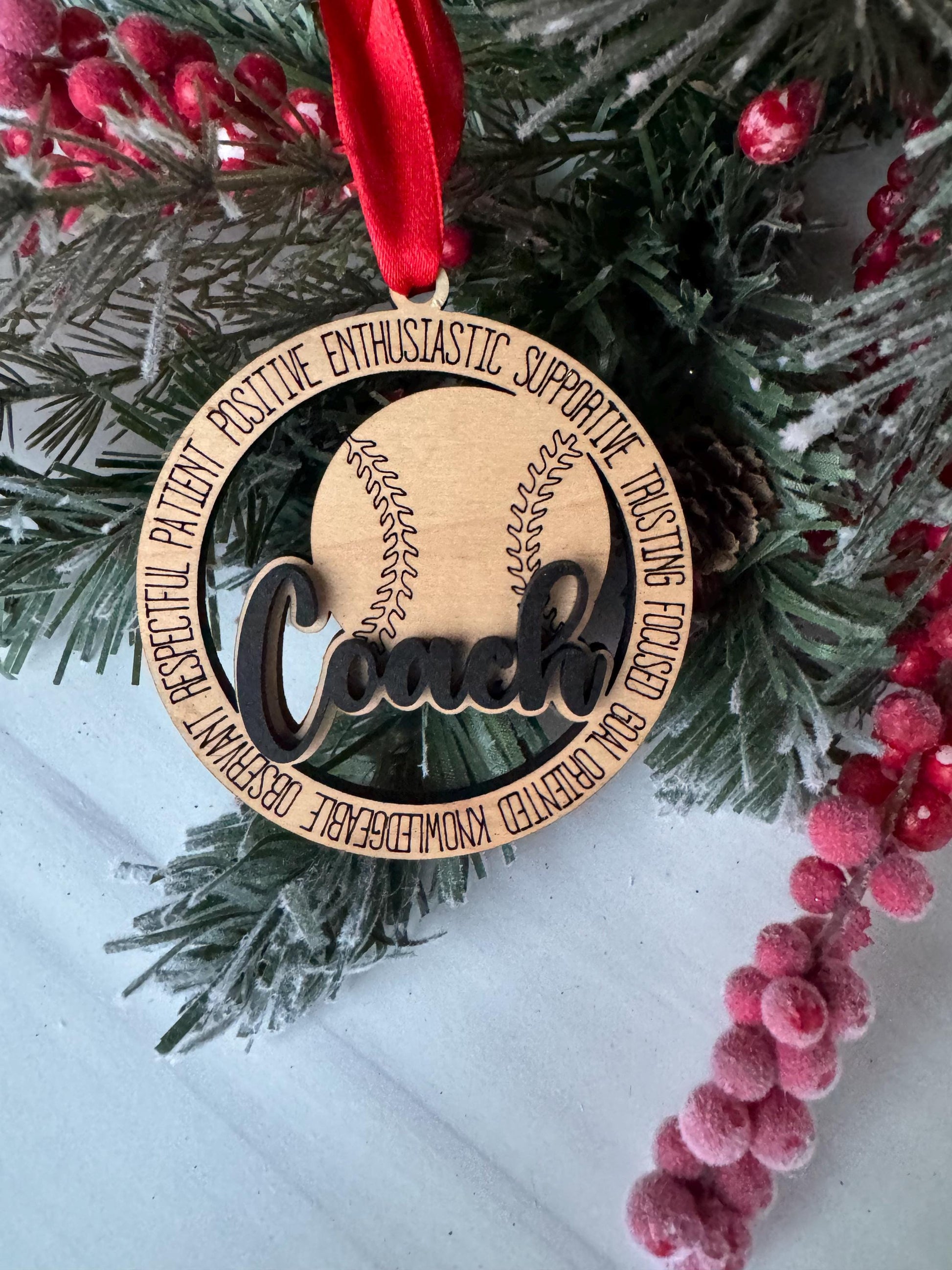 A wooden ornament with a baseball and the word "Loves" on it, placed on a pine branch with red berries.